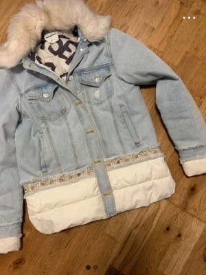 Ljusblå jeansjacka Desigual med fuskpäls - Unik ljusblå jeansjacka från Desigual med vit fuskpäls på kragen och vita quiltade detaljer nedtill och vid ärmsluten. Jackan har två bröstfickor med knappar och dekorativa sömmar. Insidan har ett coolt grafiskt mönster i svart och vitt.