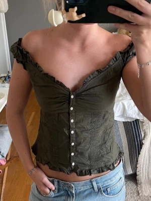 Olivgrön offshoulder topp med knappar från brandy melville - Snygg olivgrön topp med offshoulder-modell och dekorativa knappar framtill. Toppen har volangkant längs halsringningen och nederkanten, samt en croppad passform. Perfekt för dig som gillar trendiga och feminina detaljer. Har denna i Vit oxå om det är av intresse👍🏻❤️