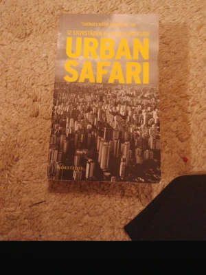 Urban Safari av Anders Karlsson - Boken 'Urban Safari' av Anders Karlsson har ett omslag med svartvit stadsbild och stora writing i g exact gul färg. Den handlar om 12 storfstäder och är utgiven av Norsted plastat pocketformat.