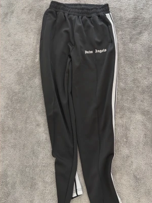 Palm Angels svarta mjukisbyxor - Palm Angels sv blanket mjukisbyxor med vita sidoränder och logus på vänster lår. Byxorna har resår i midjan och raka ben. Perfekta för en avslappnad streetwear-look.