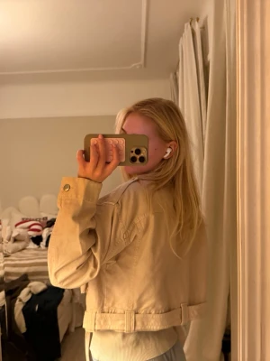 Beige/rosa cropped overshirt med fickor - Säljer en beige/rosa overshirt med cropped passform och stora fickor framtill. Jackan har knappar vid ärmslut och ett brett bälte i midjan för en cool look. Materialet är kraftigt och passar perfekt till lager-på-lager-stil.