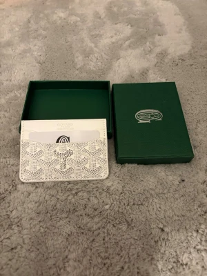 Snygg Vit korthållare - Snyggt vitt kortfodral med klassiskt mönster och flera fack för kort. Tillverkad i exklusivt läder med tryckt logotyp på framsidan. Kommer med original grön ask som ger en lyxig känsla. Perfekt för dig som vill ha stilren och exklusiv accessoar.