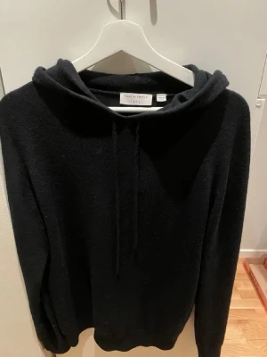 Svart hoodie från Tiger of Sweden - Mysig svart ull hoodie från Tiger of Sweden med dragsko i huvan. Tillverkad i ett mjukt och lite fluffigt ull material som känns lyxigt. Klassisk passform med långa ärmar och ribbade muddar. Perfekt för en stilren och avslappnad look.