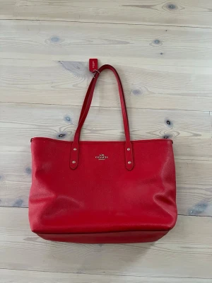 Väska från Coach - Säljer en snygg röd väska från Coach i strukturerat läderr. Väskan har silverfärgade detaljer, dubbla axelband och en Coach-tag. 