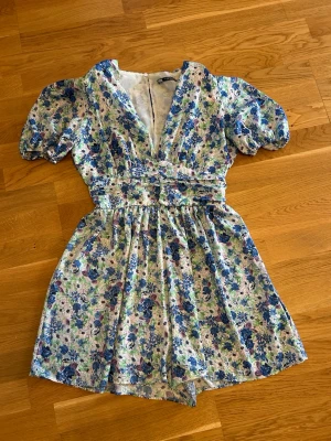  Ny Blommig jumpsuit från Zara  - Söt kortklänning med blommigt mönster i blått, grönt och lila. Klänningen har puffärmar, v-ringning och markerad midja som ger en feminin siluett. Tillverkad i mjuk bomull, perfekt för varma dagar. Passar dig som gillar romantisk och färgglad stil.