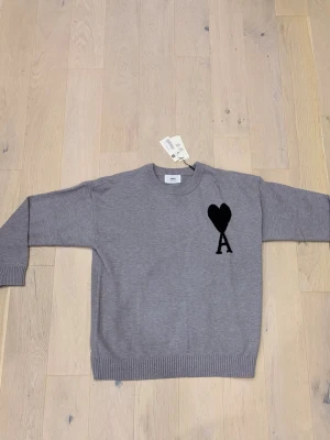 Grå stickad tröja från Ami Paris - Snygg grå stickad tröja från Ami Paris med svart hjärta och bokstaven A broderat på bröstet. Tröjan har rund halsringning, ribbade muddar vid ärmslut och nederkant samt en avslappnad passform. Perfekt för en trendig och stilren look.
