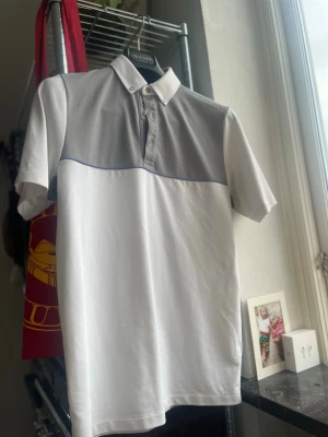 Vit och grå piké från Oscar Jacobson - Snygg pikétröja från Oscar Jacobson i vitt och ljusgrått med klassisk krage och knappar framtill. Tröjan har korta ärmar och en diskret blå linje som detalj mellan färgfälten. Perfekt för en clean och sportig stil, perfekt till golfen i sommar.