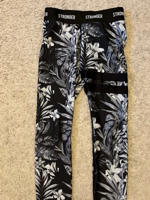 Svarta leggings med blommigt mönster Stronger - Snygga svarta leggings från Stronger med grått och vitt blommigt mönster och bred resår i midjan med logga. Tight passform och stretchigt material som passar perfekt till träning eller chill. Två svarta ränder på ena benet ger extra detalj.