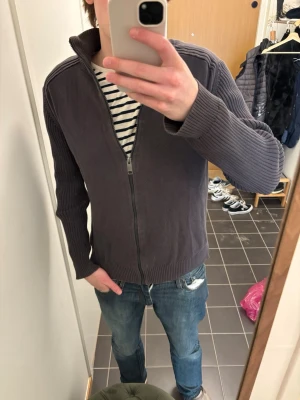 Mörkgrå stickad ziptröja - Mörkgrå stickad tröja med dragkedja framtill och hög krage. Tröjan har ribbade muddar vid ärmslut och nederkant samt en stilren, enkel design utan synliga loggor. Perfekt för lager på lager och passar till jeans eller chinos.