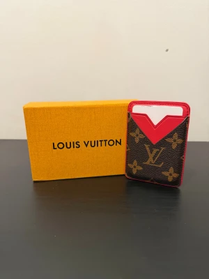 Korthållare  - Snygg och kompakt korthållare med klassiskt monogrammönster i brunt och guld, kombinerat med en stark röd detalj upptill. Tillverkad i skinn med plats för kort både fram och bak. Perfekt att fästa ( magnet) på mobilen.