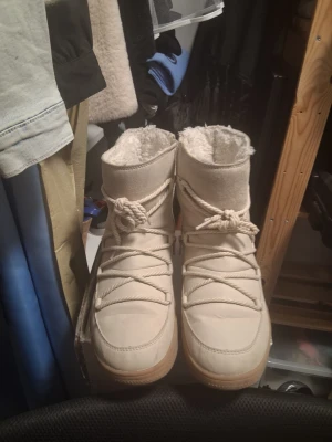 Beige moon boots med snörning - Säljer ett par beige moon boots med grov snörning och fluffigt foder. Skorna har en hög modell och rund tå, samt en ljusbrun sula. Perfekta för kalla dagar när du vill hålla värmen och ändå se stilren ut.