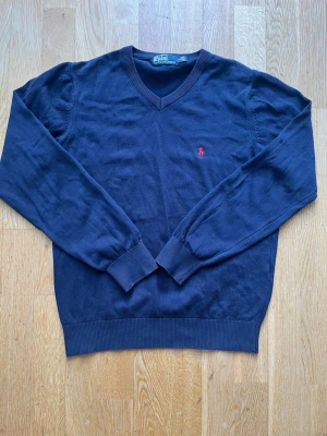 Mörkblå stickad tröja Polo Ralph Lauren - Mörkblå stickad tröja från Polo Ralph Lauren med V-ringning och röd broderad logga på bröstet. Tröjan har långa ärmar, ribbade muddar och nederkant. Perfekt till jeans eller chinos för en clean och klassisk look.