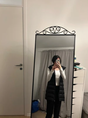 Svart lång dunväst med huva - Säljer en svart lång dunväst med huva och dragkedja framtill. Västen har ett quiltat mönster och justerbara snören vid huvan. Perfekt för lager-på-lager och ger en cool look till din outfit.