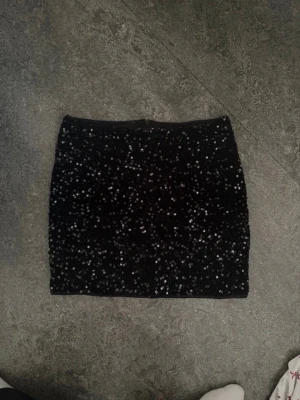 Svart paljettkjol med dragkedja - Snygg svart kortkjol täckt med glittriga paljetter. Kjolen har en smal passform och dragkedja bak för enkel påklädning. Perfekt för dig som vill sticka ut och glänsa lite extra. Materialet känns mjukt och följsamt.