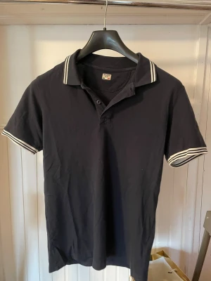 Marinblå pikétröja från Dolomite - Snygg marinblå pikétröja från Dolomite med vita ränder på krage och ärmslut. Klassisk polokrage med knappar och ett Dolomite-märke på ena ärmen. Perfekt för en sportig och stilren look. Pris kan diskuteras vid snabb affär .