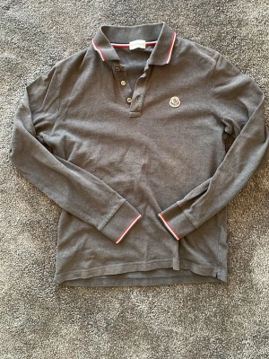 Grå långärmad piké från Moncler - Snygg grå långärmad piké från Moncler med klassisk krage och knappar framtill. Röd och vit rand på krage och ärmslut ger en sportig touch. Broderad Moncler-logga på bröstet. Tillverkad i mjuk bomull för skön känsla.