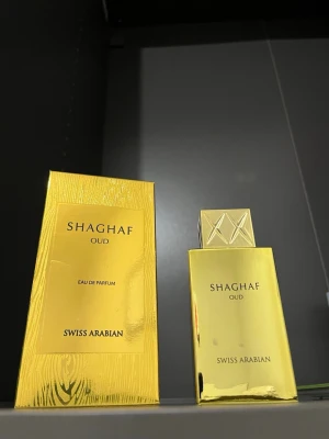 Shaghaf Oud Eau extrakt från a Swiss Arabian - Lyxig parfymflaska i guld med matchande förpackning. sopar Shaghaf Oud från Swiss Arabian. Flaskan har en elegant, rektangulär form och ett dekorativt lock med geometriskt mönster. Doften, Eau de Parfum, är känd för sina orientaliska och träiga toner.