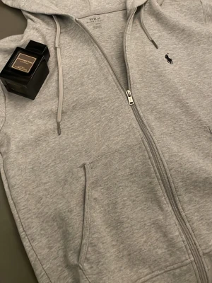 Grå zip hoodie från Polo Ralph Lauren - Säljer en ljusgrå hoodie från Polo Ralph Lauren med dragkedja, fickor framtill och justerbar huva med snören. Klassisk liten logga broderad på bröstet. Perfekt för en avslappnad och stilren look. Passar även strl M. Säljer även parfymen för 1000kr den kostar runt 2500kr ny OBS: Luvan på tröjan kan vara lite stor beroende på hur stor och lång man är.