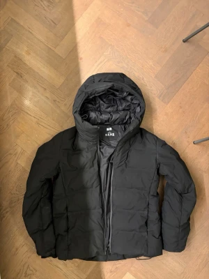 Svart pufferjacka från Uniqlo - Säljer en svart pufferjacka från Uniqlo med huva och dragkedja framtill. Jackan har en stilren design med breda kanaler och är perfekt för kalla vinterdagar. Tillverkad i ett tåligt, vattenavvisande material som håller dig varm och bekväm.