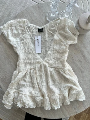 babydoll lace detail top - Supersöt vit blus från Gina Tricot med volangkant och v-ringning. Blusen har korta ärmar, smockad struktur och spetsdetaljer sopm ger en romantisk vibe. Perfekt till jeans eller kjol för en trendig look.