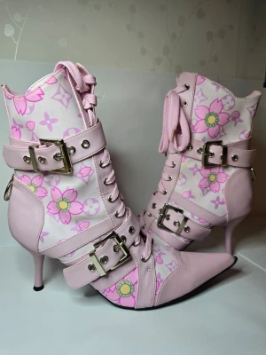 Rosa stövletter med blommönster - Unika rosa stövletter med spetsig tå, smala klackar och blommigt mönster i rosa och grönt. Skorna har snörning framtill, två breda spännen med silvriga detaljer och ringar baktill. Materialet är en mix av konstläder och textil.