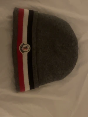 Grå Moncler mössa med ränder, helt ny har används 1 gång - Säljer en stilren grå mössa från Moncler med klassiska ränder i rött, vitt och svart längs kanten. Mössan har Moncler-loggan framtill och är tillverkad i mjukt material som sitter skönt på huvudet. Perfekt accessoar för kalla dagar.
