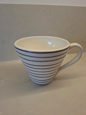 Randig kopp från Bruka Design - Säljer en vit kopp från Bruka Design med handmålade tunna lila ränder runtom. Koppen har en konisk form och ett bekvämt handtag. Märkt 'Handpainted' undertill. Modell Jumbo.