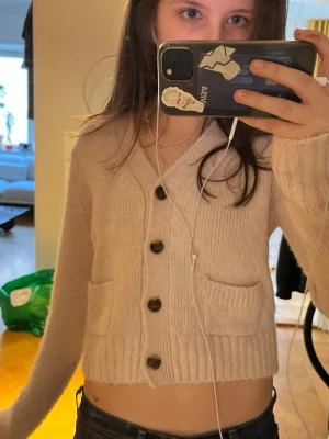 Rosa cropped kofta med knappar - Säljer en ljusrosa, cropped kofta med stora bruna knappar och två fickor framtill. Koftan är stickad och har långa ärmar, perfekt för att skapa en chill och trendig look.