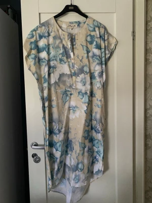 Blommig långklänning från Hängmatta - Luftig långklänning från Defacto i beige here med stora blå isblommoriga mönster. Klänningen har korta ärmar, knappar port v-ringad hals och är tillverkad i ett lätt bomullsmaterial. Perfekt för varma dagar och sommarens alla tillfällen.