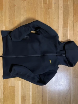 Svart Nike hoodie med dragkedja - Svart hoodie från Nike med hel dragkedja och gul Nike-logga på bröstet. Tröjan har huva och två fickor framtill. Tillverkad i mjukt och bekvämt material, perfekt för en sportig och avslappnad stil. Storlek S priset kan diskuteras 
