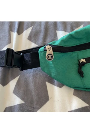 Grön magväska från Champion - Snygg grön magväska från Champion med svart justerbart band och dragkedjor. Väskan har Champion-logga på dragkedjan och på band bullet ung uptodate streetstil. Perfekt för att bära det viktigaste när du är mission on the go.