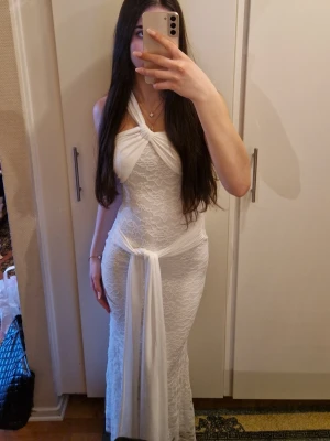 Vit långklänning med spets och omlott - Elegant vit långklänning med spetsmönster och omlottdetalj framtill. Klänningen har halterneck och en knytning vid midjan som ger en snygg siluett. Perfekt för dig som vill ha en stilren och romantisk look.