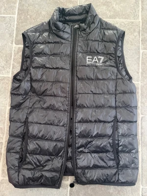 EA7 Emporio Armani väst - Snygg svart dunväst från sopiga EA7 Emporio Armani med wet-look finish och diskret logga på bröstet. Vä SAR har hög krage, drag verket framtill och två sidofickor. Perfekt för lager-på-lager och riktigt clean stil. Passar vår och sommar.