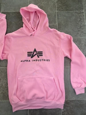 Alpha Industries Hoodie  - Säljer en snygg rosa hoodie från Alpha Industries med stor svart logga och text på bröstet. Hoodien har huva med dragsko och en stor magficka. Tillverkad i mjukt bomullsmaterial och har ribbade muddar vid ärmar och nederkant. Perfekt för en sopig streetwear-look.