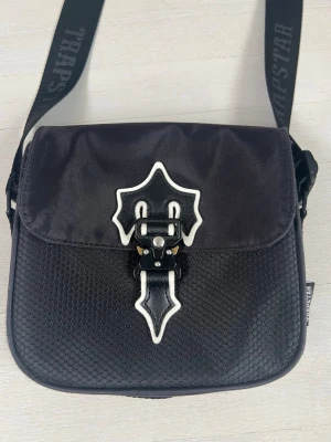 Svart axelväska från Trapstar - Snygg svart axelväska från Trapstar med justerbar axelrem och unik vitdetaljerad spännesdekor på framsidan. Väskan är tillverkad i slitstarkt tyg med bikakemönstrad struktur på framsidan och har en praktisk ficka på baksidan. Perfekt för att bära dina essentials.