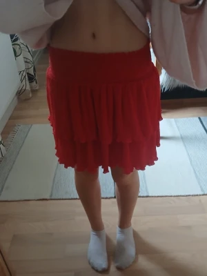 Röd volangkjol med resårmidja - Säljer en kort röd kjol med dubbla volanglager och bred resår i midjan. Kjolen har en luftig och flowy känsla, perfekt för dig som vill ha en söt och lekfull stil. Passar till både sneakers och boots.