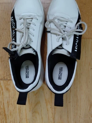Vita sneakers från Michael Kors - Snygga vita sneakers från Michael Kors med svarta detaljer och logga på sidan. Skorna har vita snören, svart hälflik och en tjock vit sula. Insidan är svartvit och materialet är slätt konstläder. Perfekta för en trendig och sportig look. Jag har i vanliga fall storlek 40 men skulle valt 39 i dessa skor, men man kan lösa det med en sula.