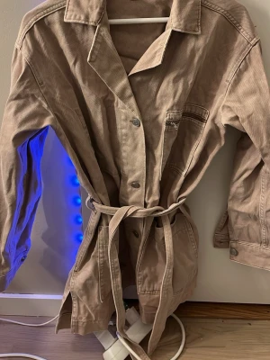 Beige trenchcoat med knytskärp - Säljer en beige trenchcoat i bomull med klassisk krage, knappar framtill och ett matchande knytskärp i midjan. Jackan har långa ärmar, fickor och en avslappnad passform. Perfekt för dig som vill ha en stilren och enkel look.