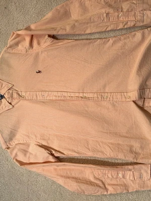 Randig skjorta från Ralph Lauren - Snygg ljusorange och vit randig skjorta från Ralph Lauren med klassisk krage och broderad logga på bröstet. Skjortan har lång ärm, knappar framtill och är i modellen Custom Fit. Perfekt för dig som gillar stilrena detaljer.