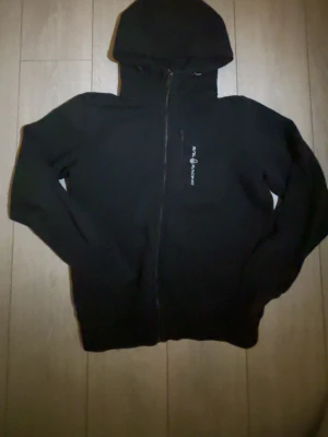 Svart zip-hoodie från Sail Racing - Svart hoodie med dragkedja från Sail Racing. Klassisk design med huva och ribbade muddar. Diskret logotyp på bröstet. Perfekt för chill dagar och streetstyle. Materialet är mjukt och bekvämt, troligen bomullsblandning.