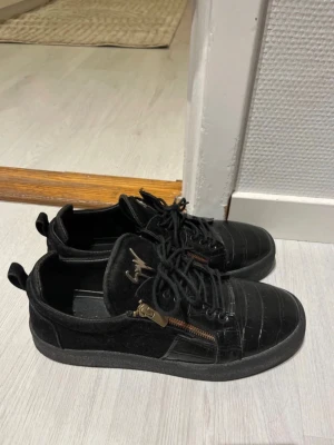 Svarta sneakers från Giuseppe Zanotti - Säljer ett par svarta sneakers från Giuseppe Zanotti med coola guldfärgade dragkedjor på sidorna och läderdetaljer. Skorna har låg profil, snörning och logotyp på tungan. Perfekt för dig som vill ha en stilren och edgy look.