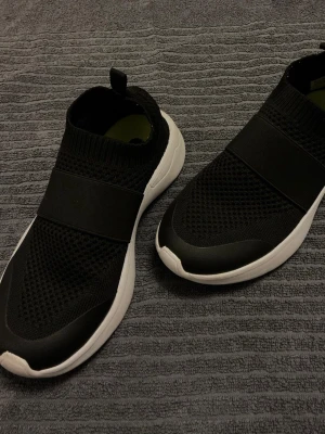 Svarta slip-on sneakers med vit sula, barn storlek 32 - Snygga svarta slip-on sneakers med stickad ovandel12 och bred resår över foten. Skorna har en vit, chunky sula och en liten flärp bak för enkel påtagning. Ovandelen är i