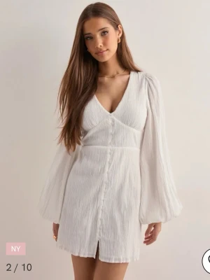 Vit kortklänning med v-ringning - Vit kortklänning med v-ringning och knappar framtill. Klänningen har långa, luftiga ärmar och en lätt struktur i tyget. Perfekt för sommarens alla tillfällen och ger en fräsch look.