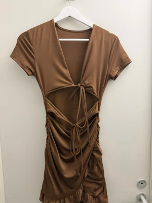 Brun kort klänning med snörning - Trendig brun kortklänning med djup v-ringning, cut-out och snörningsdetaljer framtill. Klänningen har korta ärmar, rynkade sidor och volangkant nedtill. Tillverkad i ett mjukt, stretchigt syntetmaterial som ger en slim passform.