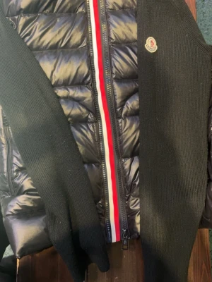 Svart cardiganjacka från Moncler - Snygg svart cardiganjacka från Moncler med quiltad front i blankt material och stickade ärmar. Dragkedja framtill med röd, vit och blå rand längs kanten. Moncler-logga på bröstet. Perfekt för dig som gillar exklusiv streetwear.