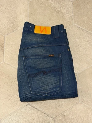 Nudie jeans - Snygga mörkblå jeans från Nudie Jeans med klassisk femficksdesign och orange logotyppatch bak. Jeansen har en slim fit-modell och är tillverkade i denim med lätt slitning och kontrastsömmar. Perfekta för en trendig och avslappnad look. Vid minsta intresse, kontakta då mig!⭐️