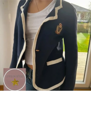 Marinblå kavaj med vita detaljer - Stilren marinblå kavaj med vita kontrastkanter och broderad emblem på bröstet. Kavajen har en knapp framtill, två fickor och är figursydd för en snygg siluett. Perfekt för en preppy look och passar till jeans eller kjol.