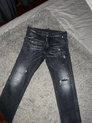 Dsquared2 Jeans - Säljer ett par grå jeans från Dsquared2 med slitningar och färgstänk för en stökig, trendig look. Jeansen har smal passform, klassiska fem fickor och detaljerade sömmar. Perfekta för dig som gillar streetwear och vill sticka ut.