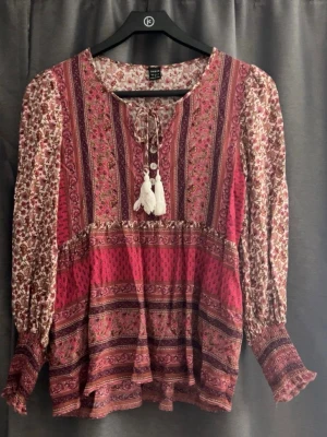 Bohemisk mönstrad blus  - Bohemisk blus i vinrött och rosa med blommigt och etnoinspirerat mönster. Långärmad modell med resår vid ärmslut, rund halsringning med knytband och tofsar. Tillverkad i ett lätt, vävt material som ger ett flowy fall.