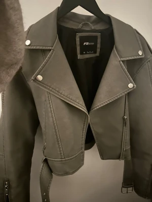 Grå skinnjacka från F.A Sister - Snygg grå skinnjacka från F.A Sister med klassisk bikerkrage, silverfärgade dragkedjor och knappar. Kort modell med bälte i midjan och coola detaljer som ger en edgy vibe. Perfekt för att lyfta vilken outfit som helst.